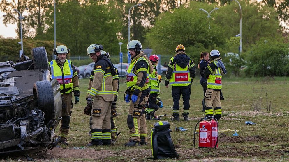 Räddningspersonal arbetar vid en bil som voltat och hamnat upp och ner på en gräsplan.