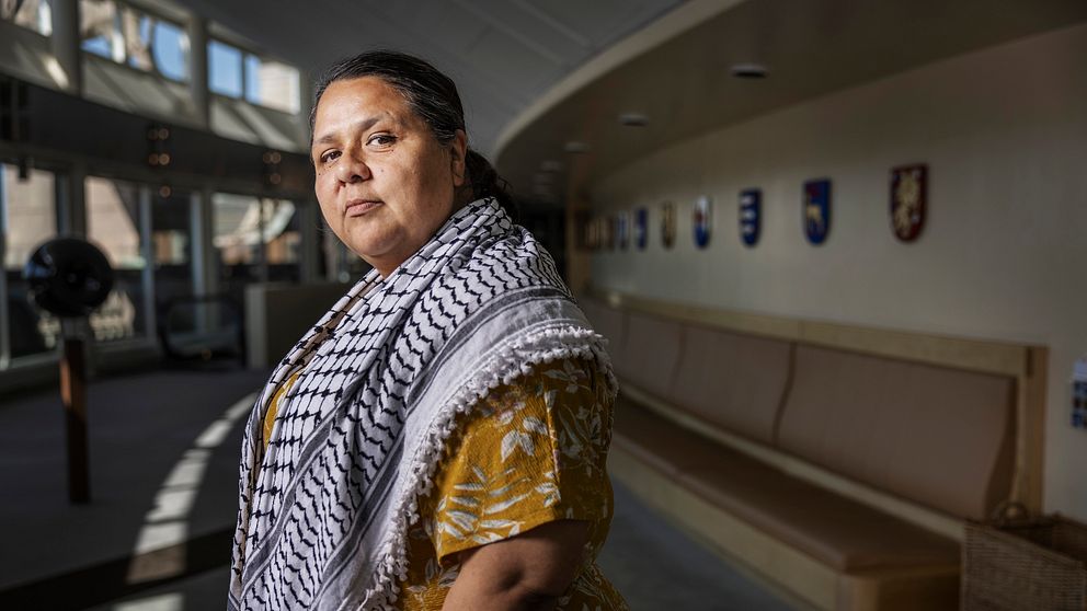 Person i gul mönstrad tröja och sjal står i en ljus hall med bänkar och sköldar på väggen.