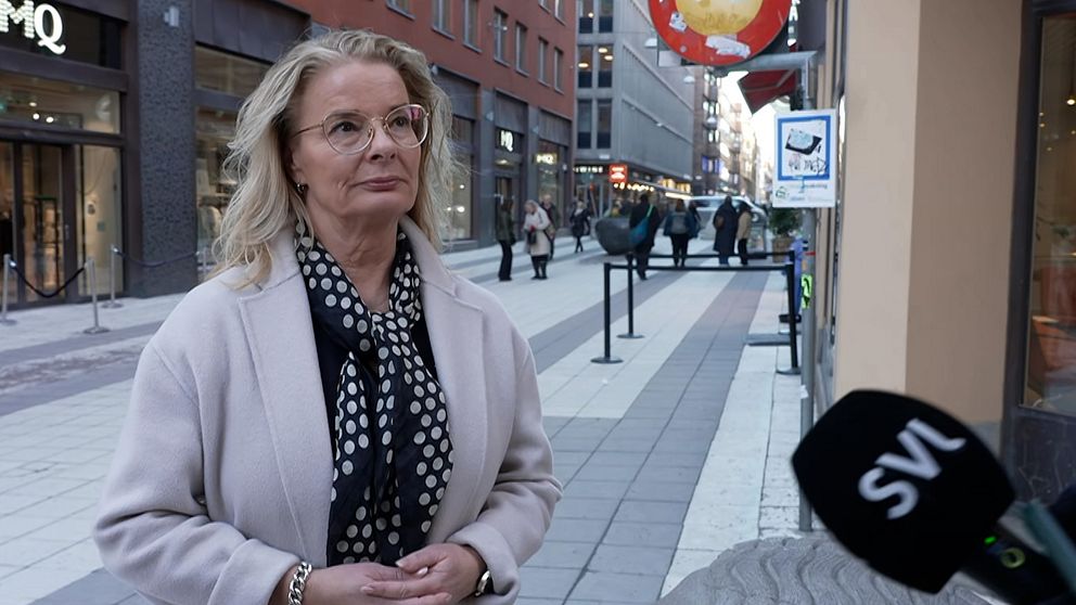 Kvinna klädd i ljus kappa och prickig halsduk står på en gågata i stadsmiljö, medan en mikrofon med ”SVT”-märke är riktad mot henne, vilket indikerar en pågående intervju.