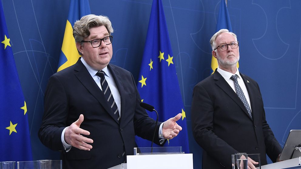 Två personer i kostym talar vid podier framför svenska och EU-flaggor på en presskonferens.