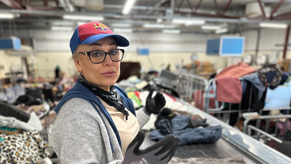 Madlin Benjamin står vid ett sorteringsband i en anläggning fylld med kläder och textilier. Personen bär en keps, handskar och en scarf samt en tröja. Runt omkring syns högar av kläder i olika färger och mönster, samt skyltar och infrastruktur från anläggningen.