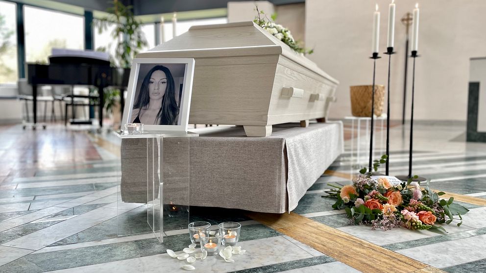 En kista placerad i ett ljust rum, omgiven av ett blommarrangemang och ljus. Framför kistan står en ram med ett fotografi, och på golvet ligger några tända ljus.