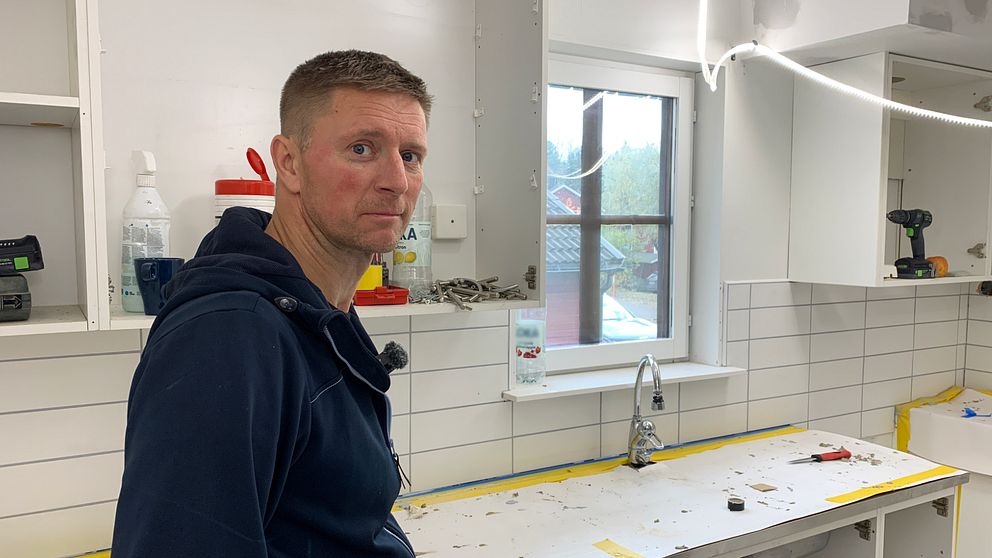 En person står vid en köksbänk i ett hem, omgiven av en arbetsyta med verktyg och material. Rummet har vita väggar och ett fönster som släpper in naturligt ljus. Det finns hyllor med rengöringsmedel och verktyg i bakgrunden.