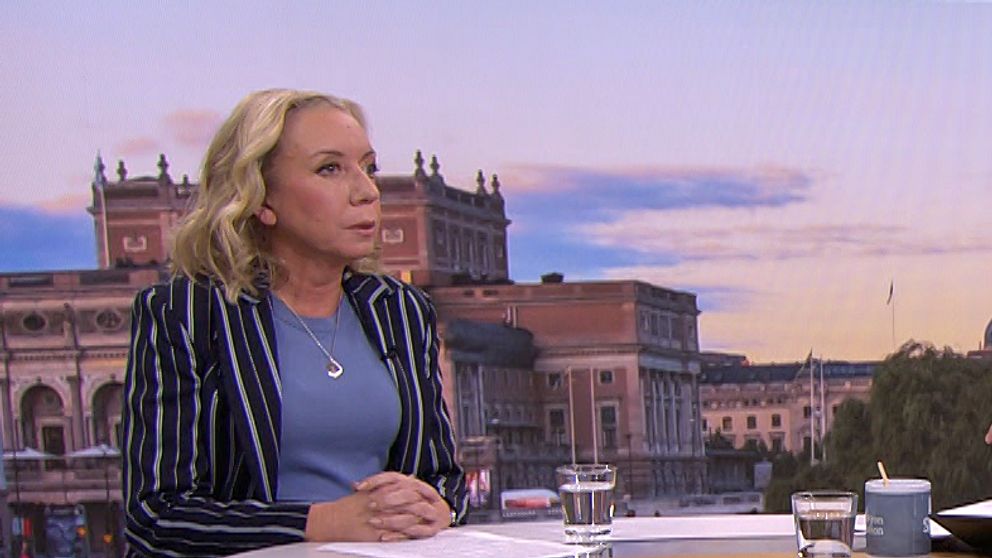 Tre personer sitter vid ett rundat bord i en TV-studio. Bakom dem syns en stadsskyline med byggnader och en färgglad himmel, vilket antyder att det är morgon eller sen eftermiddag. De två personerna som är vända mot kameran diskuterar medan den tredje personen, som sitter snett mot kameran, lyssnar. På bordet finns kaffemuggar och surfplattor.