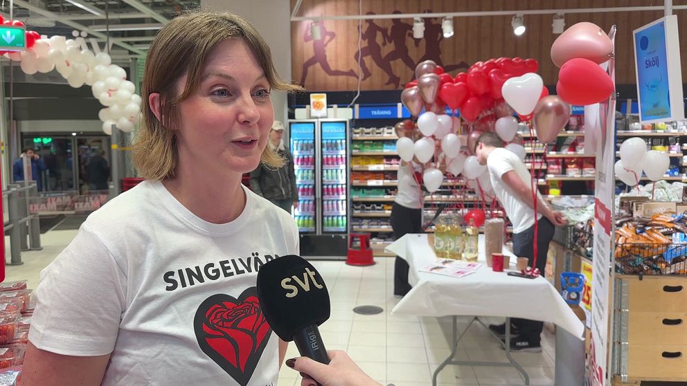 En kvinna bär en t-shirt med texten ”SINGELVÄRD” och står inför en kamera i en butik. I bakgrunden syns en monter med röda och vita ballonger samt dekorationer i samband med ett evenemang. Det finns flera personer som interagerar i butiken, vilket ger en livlig atmosfär.