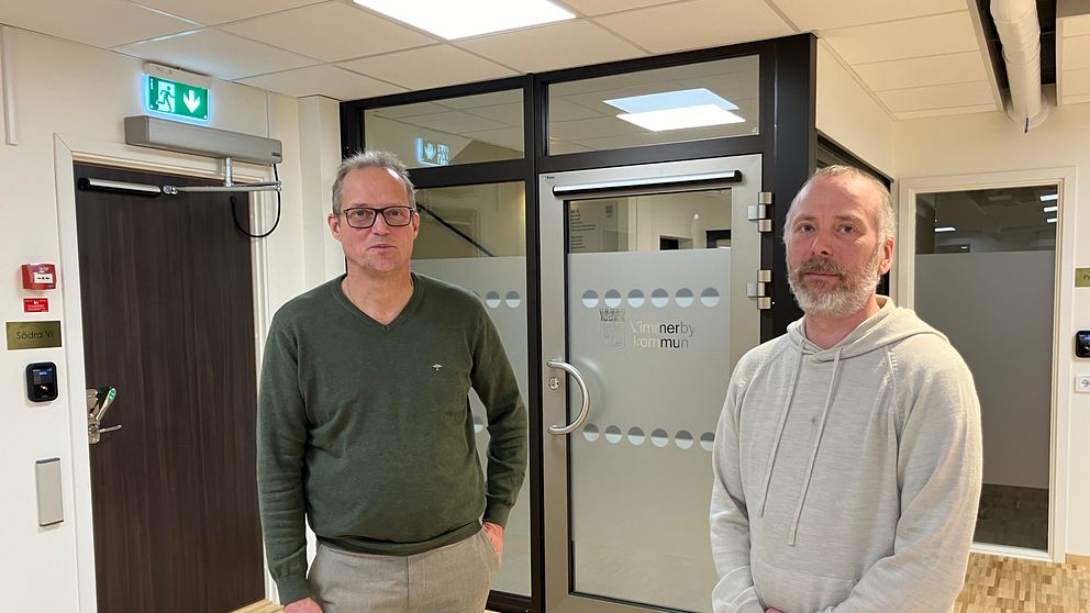 Två personer står i en korridor framför en glasdörr. Den första personen bär en grön tröja och ljusa byxor, medan den andra har en ljusgrå sweatshirt på sig. Korridoren har en modern inredning med trägolv och vita väggar. Bakom dem finns en dörr med dekaler som indikerar en kontorsmiljö.