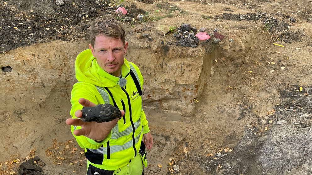 Arkeolog håller upp stenyxa av porfyr i grop vid utgrävning