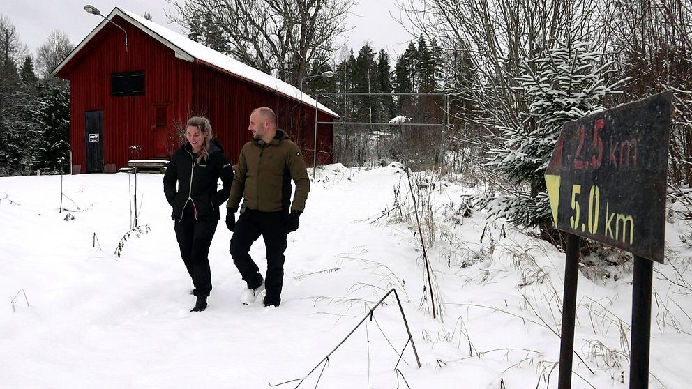 En kvinna och en man går vinterklädda längs en skogsstig med mycket snö på och runt. I bakgrunden syns en trälada.