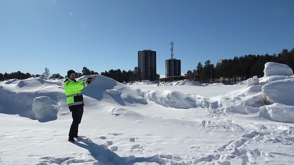 Projektledaren Erik Johansson visar var Kronans matbutik ska finnas – står i varselkläder på en snötäckt yta med två höga hus och skog i bakgrunden