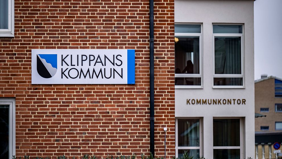 Skylt med texten ”KLIPPANS KOMMUN” på en tegelvägg till vänster, och en byggnad med fönster som har texten ”KOMMUNKONTOR” på väggen till höger. Grön buskage syns i förgrunden.