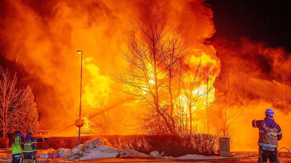 Brandmän arbetar på platsen för en massiv byggnadsbrand under natten, där intensivt orangea lågor och tjock rök stiger mot himlen. Träd och snö ses i förgrunden medan elden lyser upp omgivningen.