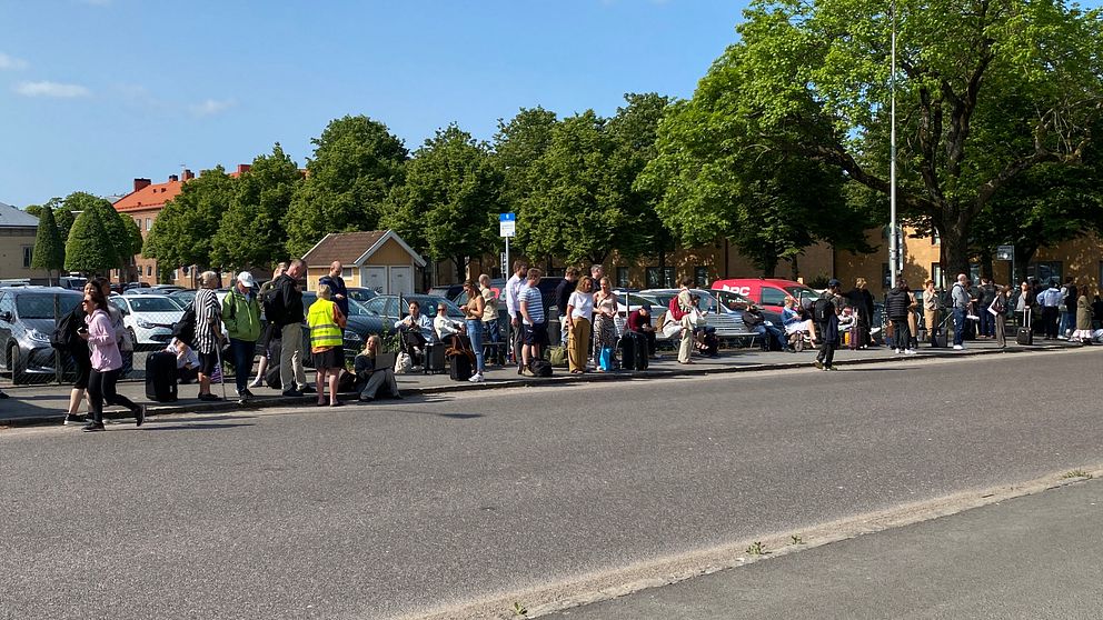 Människor i kö vid busshållplats. Gata i förgrunden.