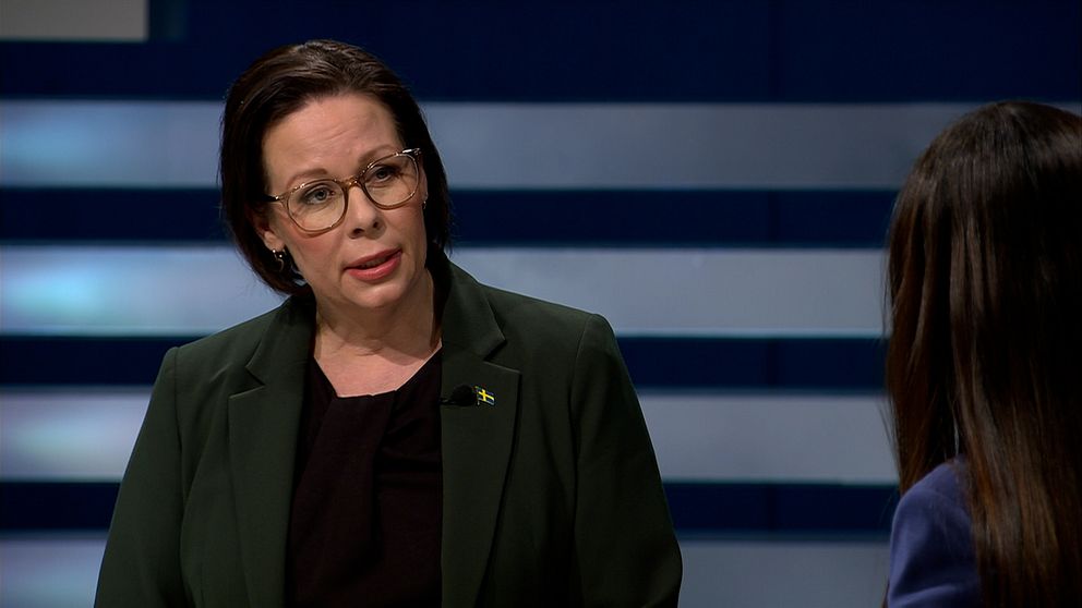 En kvinna i en grön kavaj står framför en mörk bakgrund med horisontella linjer. Hon samtalar med en annan person vid sidan av, som är delvis synlig. Kvinnan i kavajen har ett svenskt flagga-badge på sin klädd och visar en professionell attityd under diskussionen.