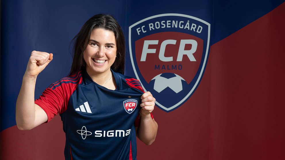 En kvinna i fotbollströja från FC Rosengård poserar med knuten näve framför sin bröst. I bakgrunden syns klubbens logotyp och färger.