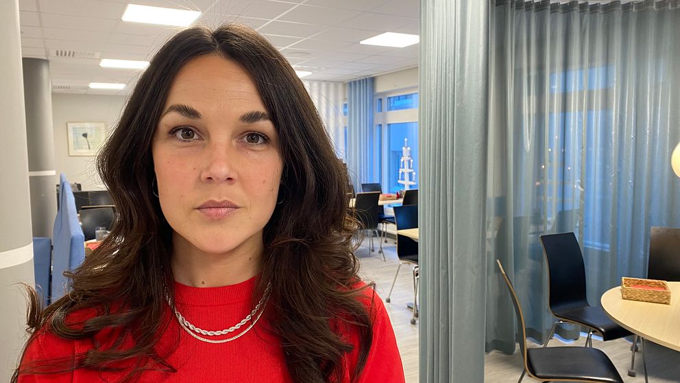 En person i en röd tröja står i ett kontorslignande utrymme, med en modern inredning och flera stolar och bord synliga i bakgrunden. Rummet har stora fönster som släpper in naturligt ljus och är avskilt med mjuka fördragna gardiner.