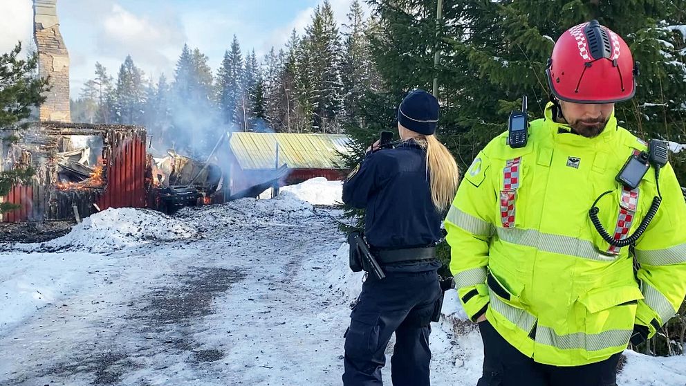 Brandman och polis vid utbränd byggnad i snöigt landskap. Rök stiger från ruinerna.