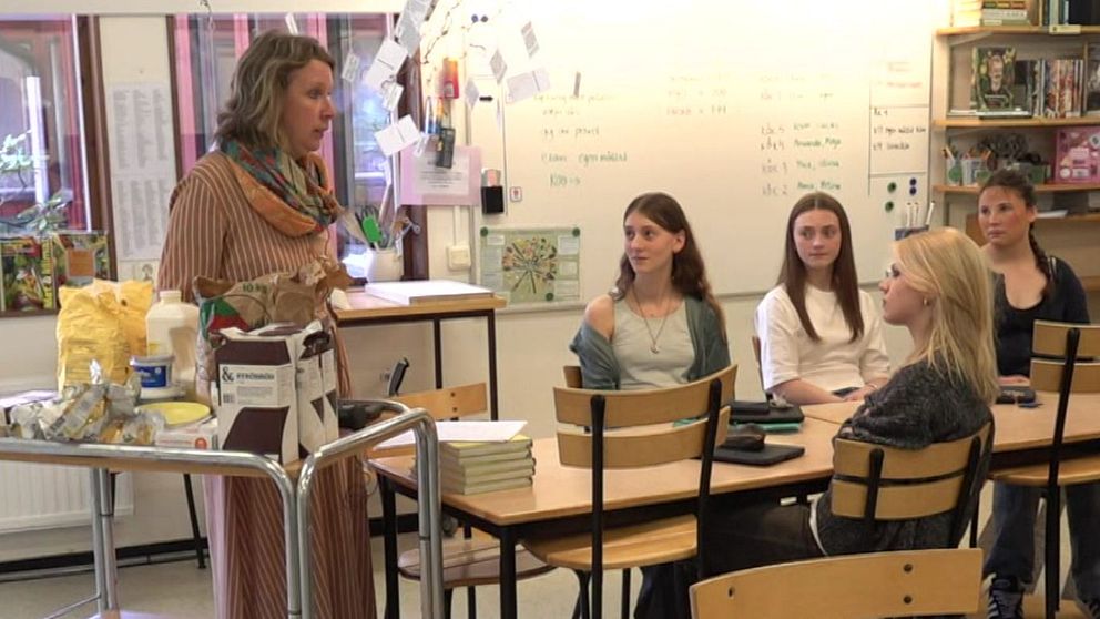 Lärare håller lektion för en grupp elever i klassrum. Elever sitter vid sina bänkar.