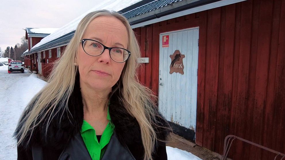 En kvinna med ljust hår och glasögon står framför ett rött hus med en skylt där det står MRK och är en häst på.