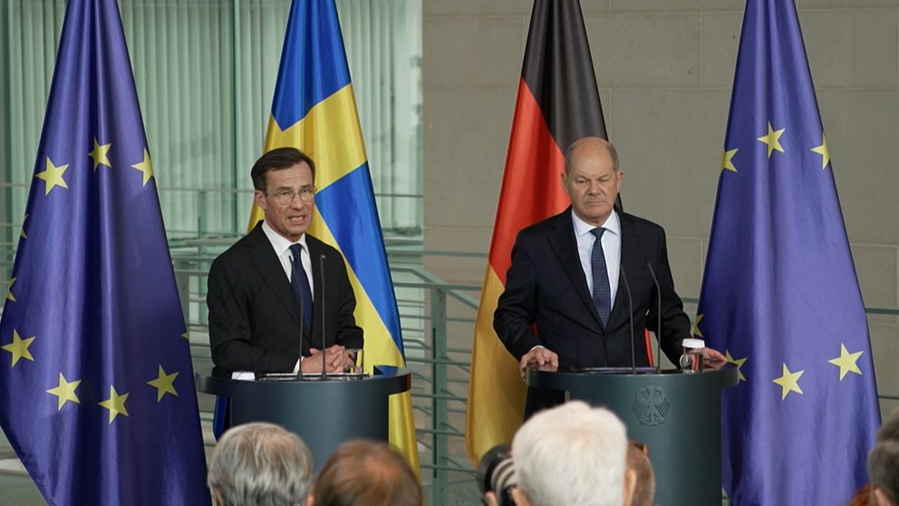 Presskonferens med två talare vid podium, där den svenska flaggan och den tyska flaggan är synliga. Bakom dem står EU-flaggor, vilket understryker det europeiska samarbetet.