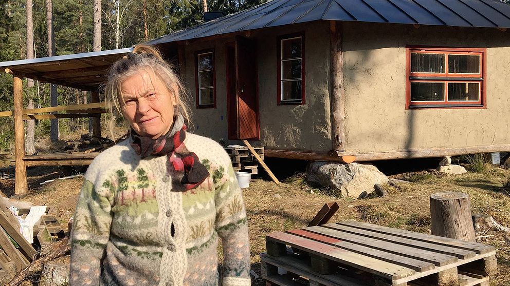 Barbro i stickad kofta står framför det lerfärgade huset med röda fönster.