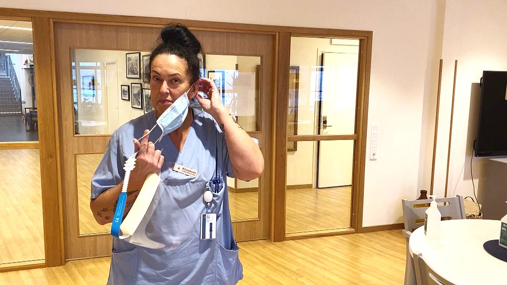 En sköterska som står framför några dörrar, och tar på sig munskydd. Håller visir i handen.