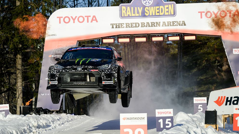 En rallybil svävar i luften efter ett hopp under Rally Sweden vid The Red Barn Arena. Omkringliggande snö och träd indikerar vinterförhållanden. Skyltar markerar sträcknummer och hastighet.
