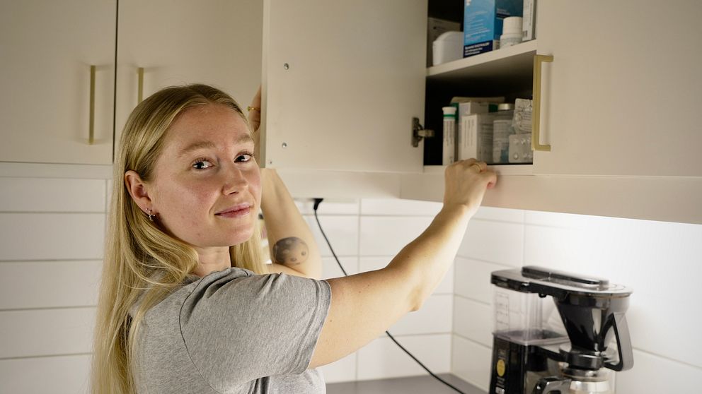 En person med långt blont hår står i ett kök och plockar fram föremål från ett överskåp. På den grå köksbänken nedanför står en kaffebryggare.