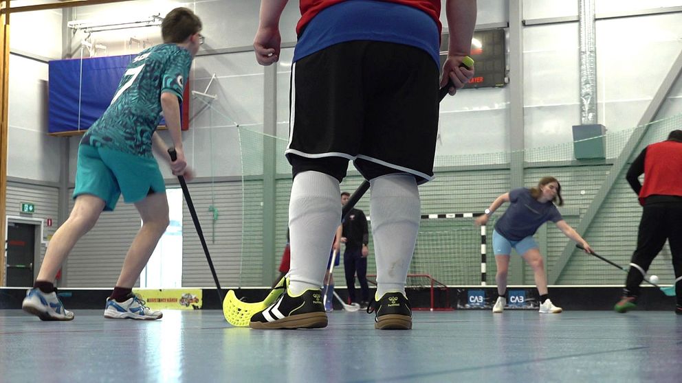 Unga spelare spelar innebandy i en idrottshall. Klubbfäktning nära målet.