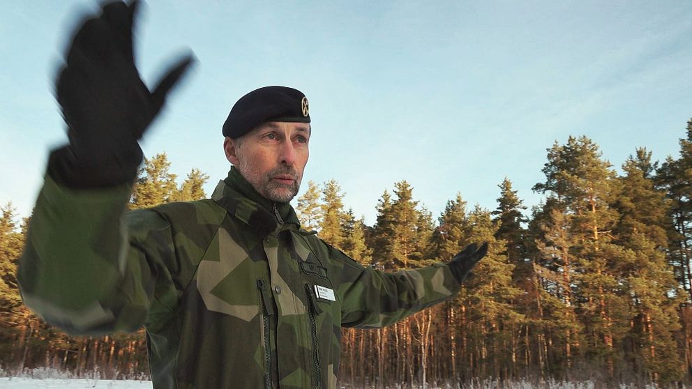 Man i militärkläder gestikulerar med armarna framför blå himmel och skog