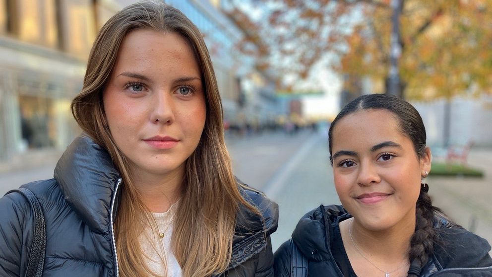 Två personer står tillsammans utomhus, iförda svarta jackor, på gågata i Uppsala med träd och byggnader i bakgrunden.