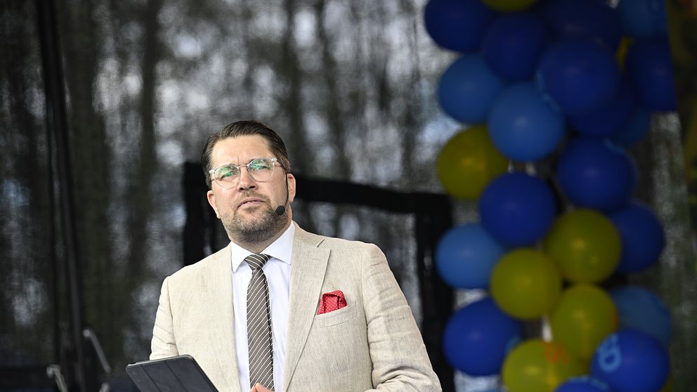 Man i beige kavaj talar på scen bredvid blå och gula ballonger.