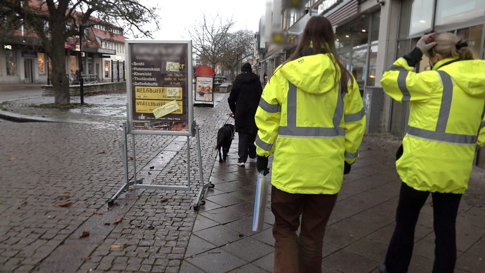 Två kvinnor i gula jackor tittar på skyltar som står på gatan i visby.