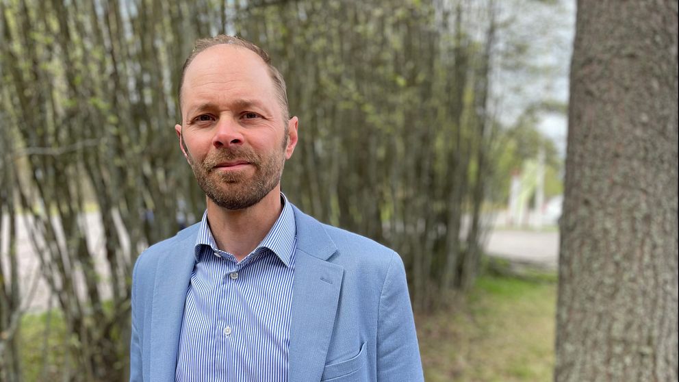 Man i ljusblå kavaj står utomhus vid träd och buskar.