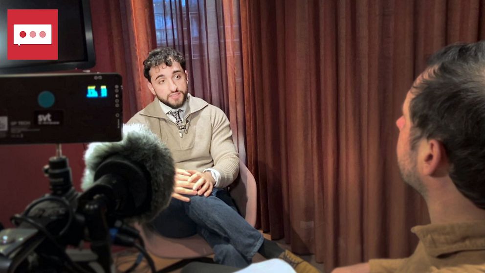 Intervjuperson i fåtölj pratar med journalist i ett rum med mörka gardiner, filmutrustning synlig.