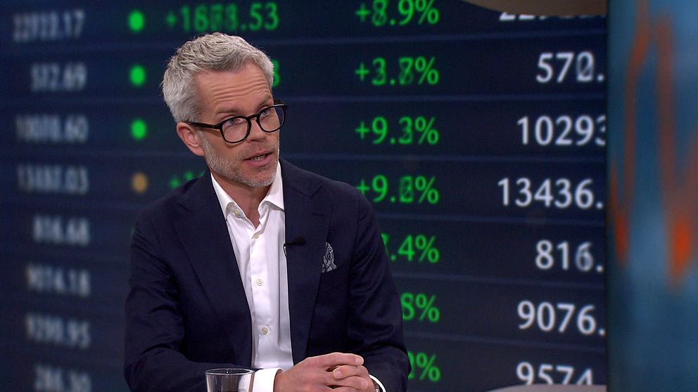 Person i kostym i tv-studio med börskurser och siffror synliga i bakgrunden.