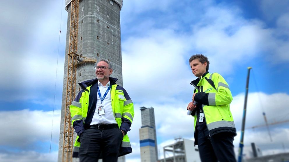 Två personer i gula säkerhetsjackor står i förgrunden, diskuterande. I bakgrunden syns en hög byggnad under konstruktion, prydd med byggnadsställningar och en kran. Himlen är delvis molnig.