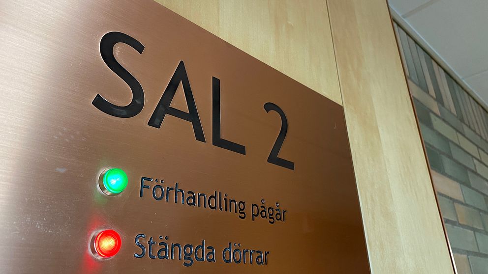En skylt utanför en tingsrättssal i Nyköping. På skylten står det ”Sal 2”. Under det står det ”Förhandling pågår”, där det lyser en grön lampa bredvid. Under det står det ”Stängda dörrar” där det lyser en röd lampa bredvid.