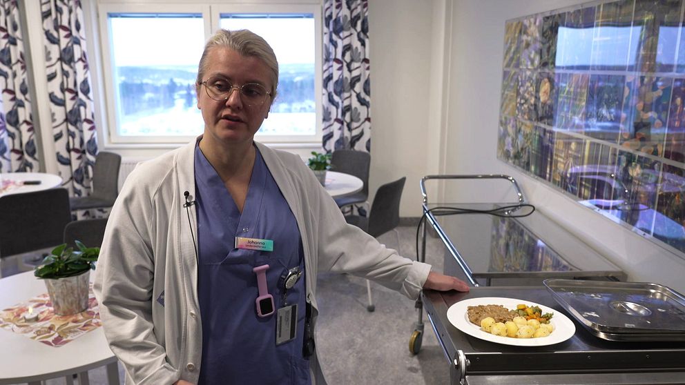 En person i vårdkläder står vid en serveringsvagn inomhus, bredvid en tallrik med mat som innehåller köttfärs och grönsaker. I bakgrunden syns fönster med utsikt samt bord och stolar i en ljus och modern miljö. Det finns också en växt på bordet och en färgglad väggdekoration i rummet.