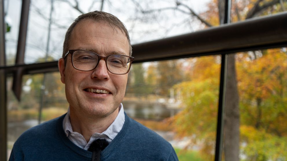 Regionstyrelsens ordförande Patrik Stenvard (M) står i fokus med bakgrund av en grön och orange höstlandskap genom ett stort fönster. Ljuset är mjukt och naturen syns tydligt, vilket skapar en lugn atmosfär.