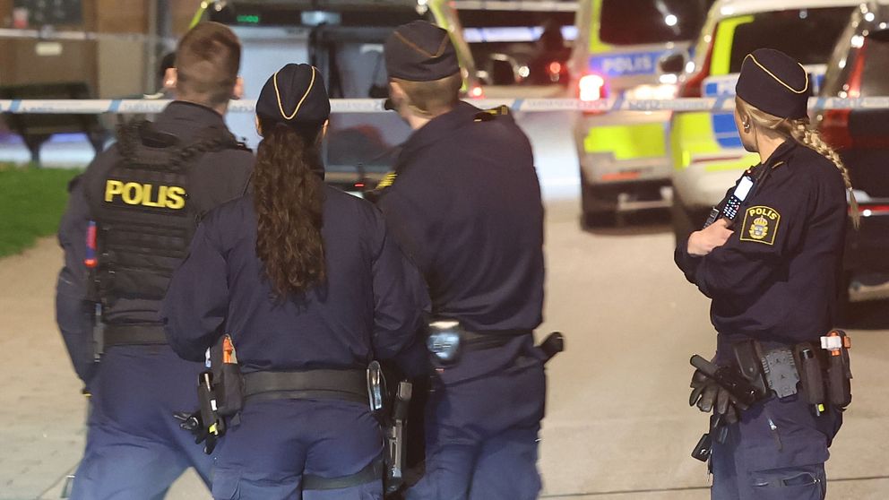 Fyra poliser står vid avspärrning, polisfordon med blåljus syns i bakgrunden.