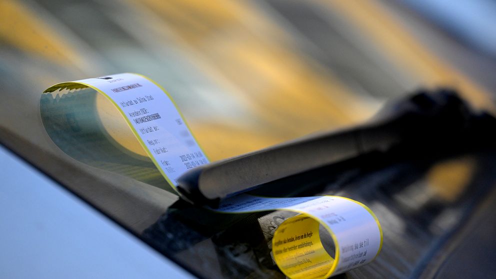 Parkeringsbot fäst på vindrutetorkaren av en bil, med rullen av betalningskvitton delvis synlig.
