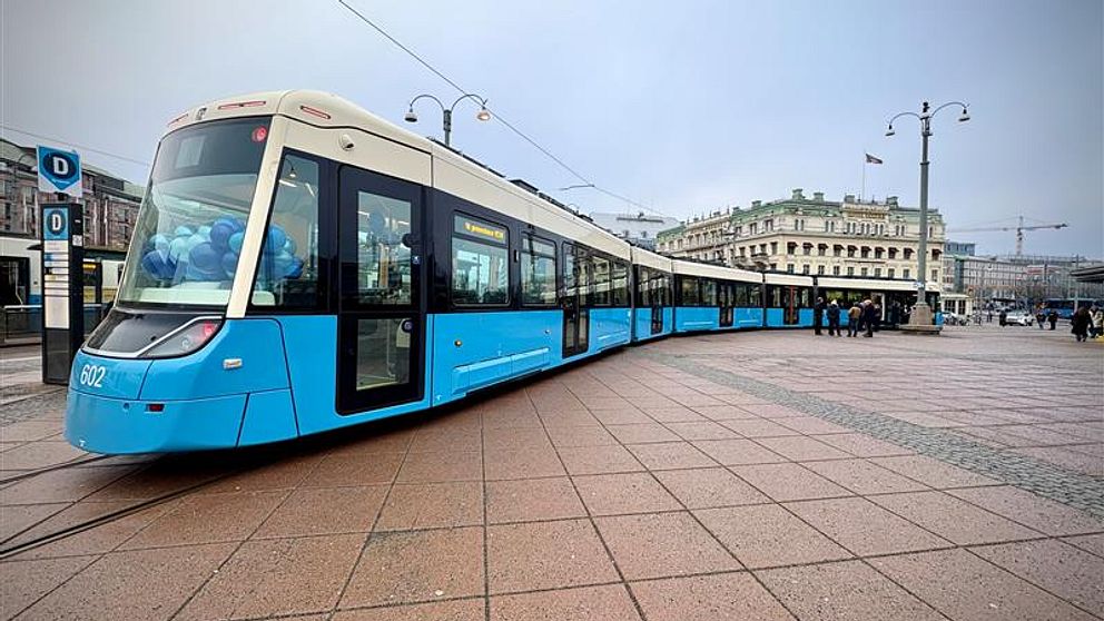En modern spårvagn i ljusblå färg står på en torgyta med stora plattor. I bakgrunden syns historisk arkitektur och några personer som går förbi, med en lätt dimma över området.
