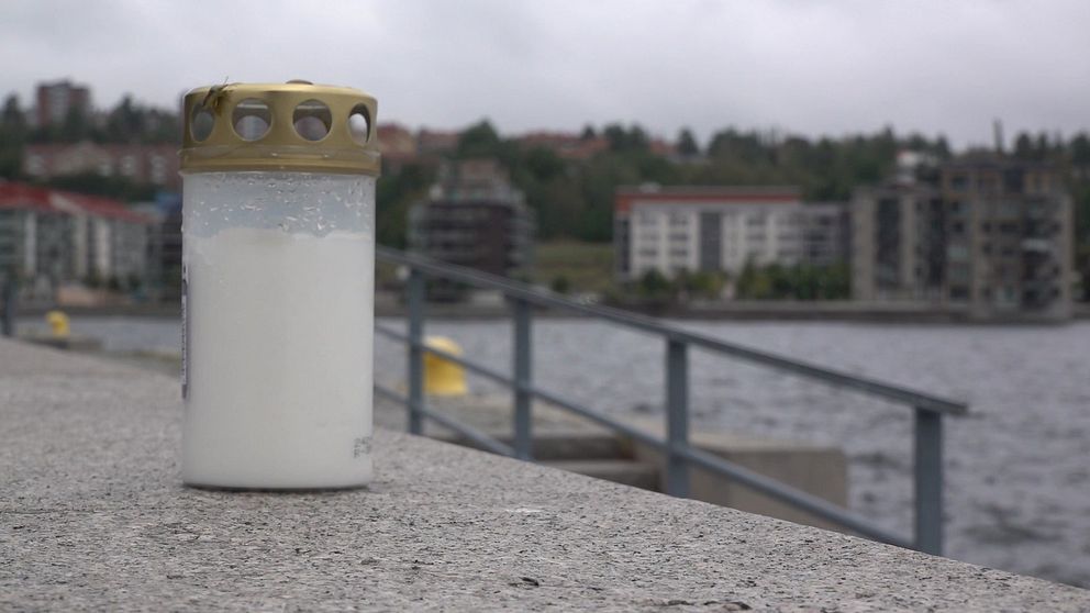 Ett vitt gravljus med ett guldlock står på kajkanten vid vattnet i Sundsvalls hamn. I bakgrunden syns byggnader och en molnig himmel, vilket skapar en lugn men dyster atmosfär.