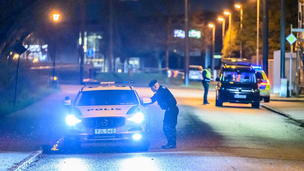 En polisman står vid en polisbil med påslagna ljus på en mörk gata. I bakgrunden syns en annan polisbil och poliser som arbetar i området. Gatubelysning och omgivande miljö ger en känsla av nattlig övervakning.