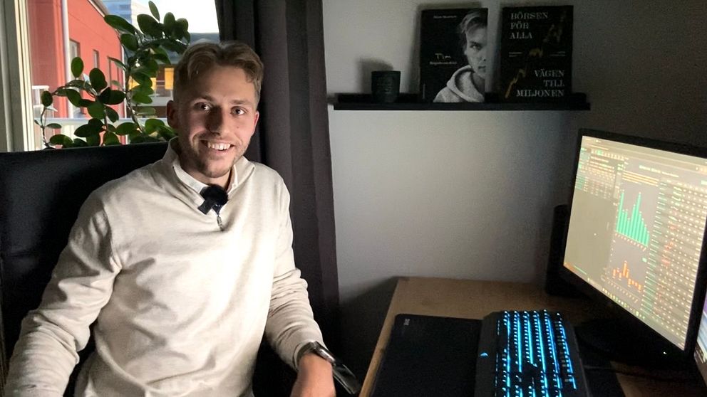 Oliver Berg sitter vid ett skrivbord i ett kontorsutrymme, klädd i en ljus tröja och skjorta. På skrivbordet finns en dator med en färgglad skärm som visar en graf eller diagram, och i bakgrunden syns en växt och hyllor med böcker eller dekorationer. Rummet har fönster som släpper in ett svagt ljus.