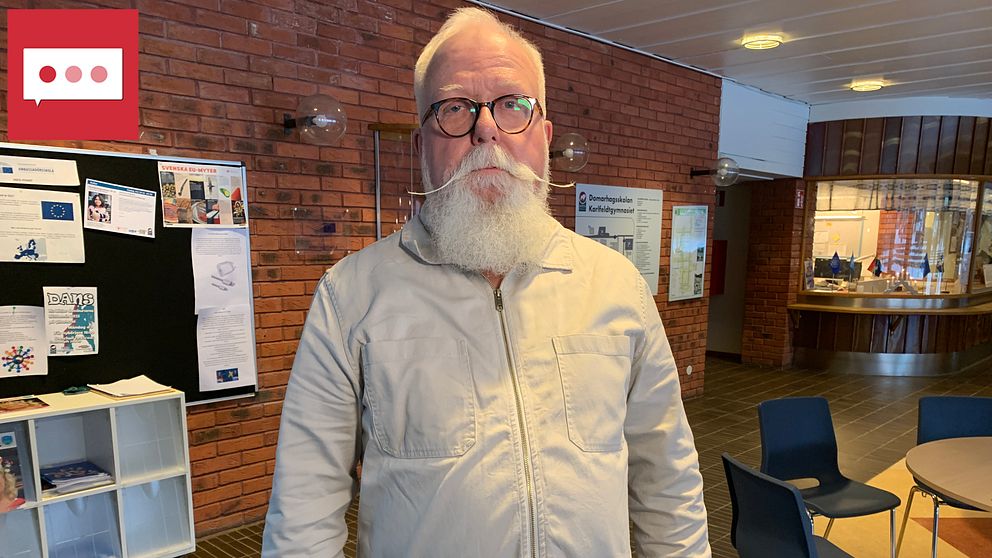En man med lång spetsig mustasch tittar in i kameran i en skolcafeteria.