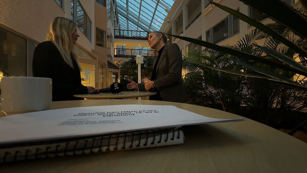 Två personer står och samtalar i en ljus, växtfylld atrium, med en stor glasad takstruktur. I förgrunden syns en anteckningsbok och en mugg, vilket tyder på att de diskuterar ett arbetsrelaterat ämne.