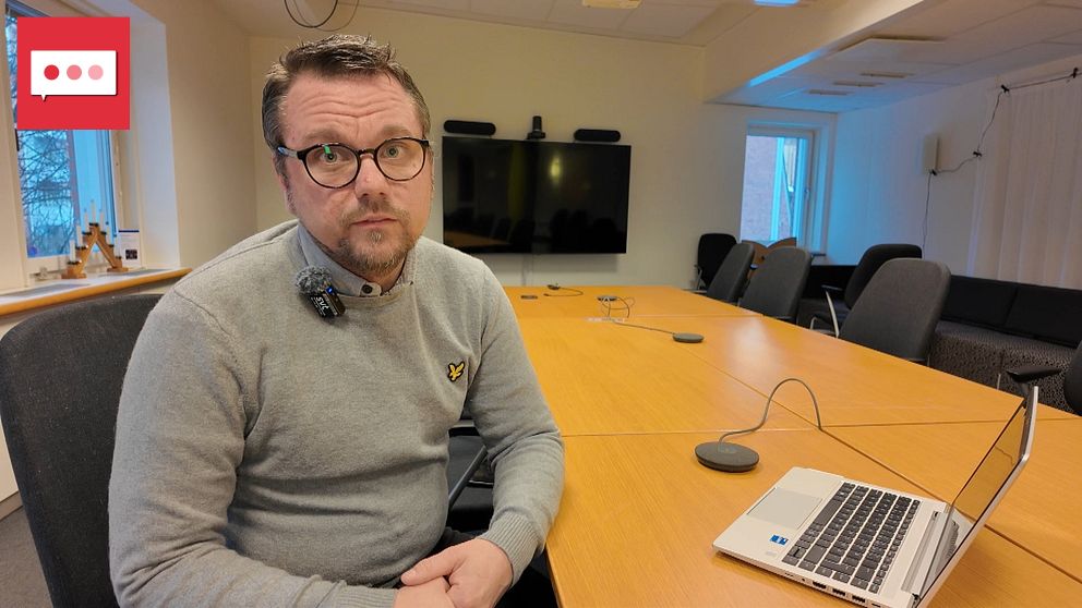 En person sitter vid ett konferensbord i ett rum med modern inredning. Framför honom står en laptop och på bordet syns mikrofon och mötesutrustning. I bakgrunden finns en stor skärm. Rummet är ljust och har stora fönster som ger insyn till utomhusmiljön.