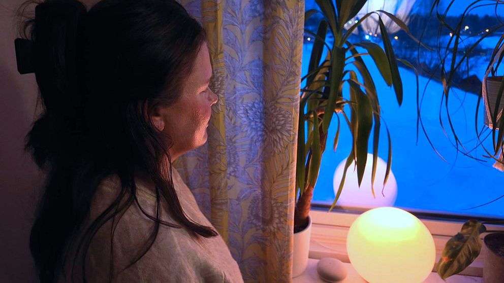 Petra Alfredsson står vid ett fönster på kvällen, belyst av en varm glödlampa. Gardiner med blomstertryck och en grön växt bredvid fönstret.
