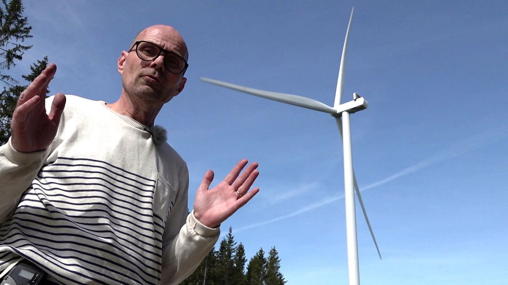 Person i randig tröja framför vindkraftverk mot blå himmel och skogskant.